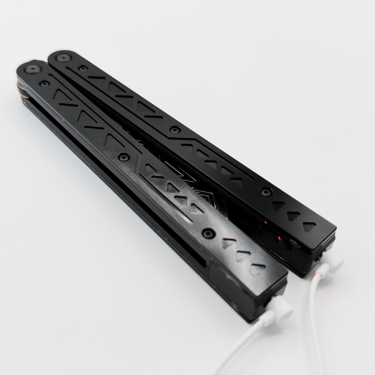 Black balisong trainer with intricate cutout designs on the handles, resting closed on a white background with charging cables attached.