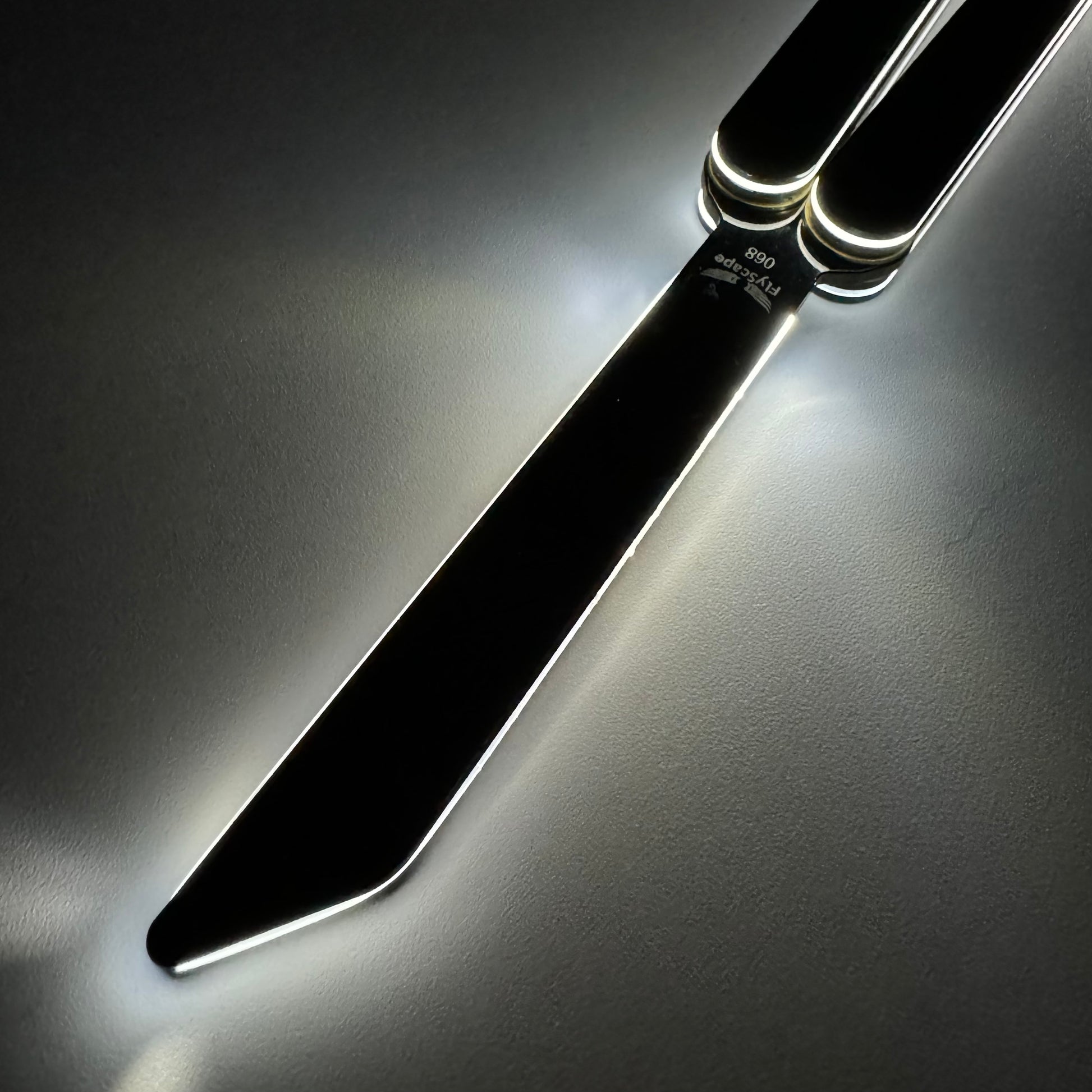 A close-up of a black butterfly knife, also known as a balisong or butterfly trainer, with its blade fully extended. The knife, also referred to as a Sabre, is photographed in low light, with glowing edges reflecting off the polished metal surface. The dark background and soft lighting emphasize the knife’s modern and sleek design.