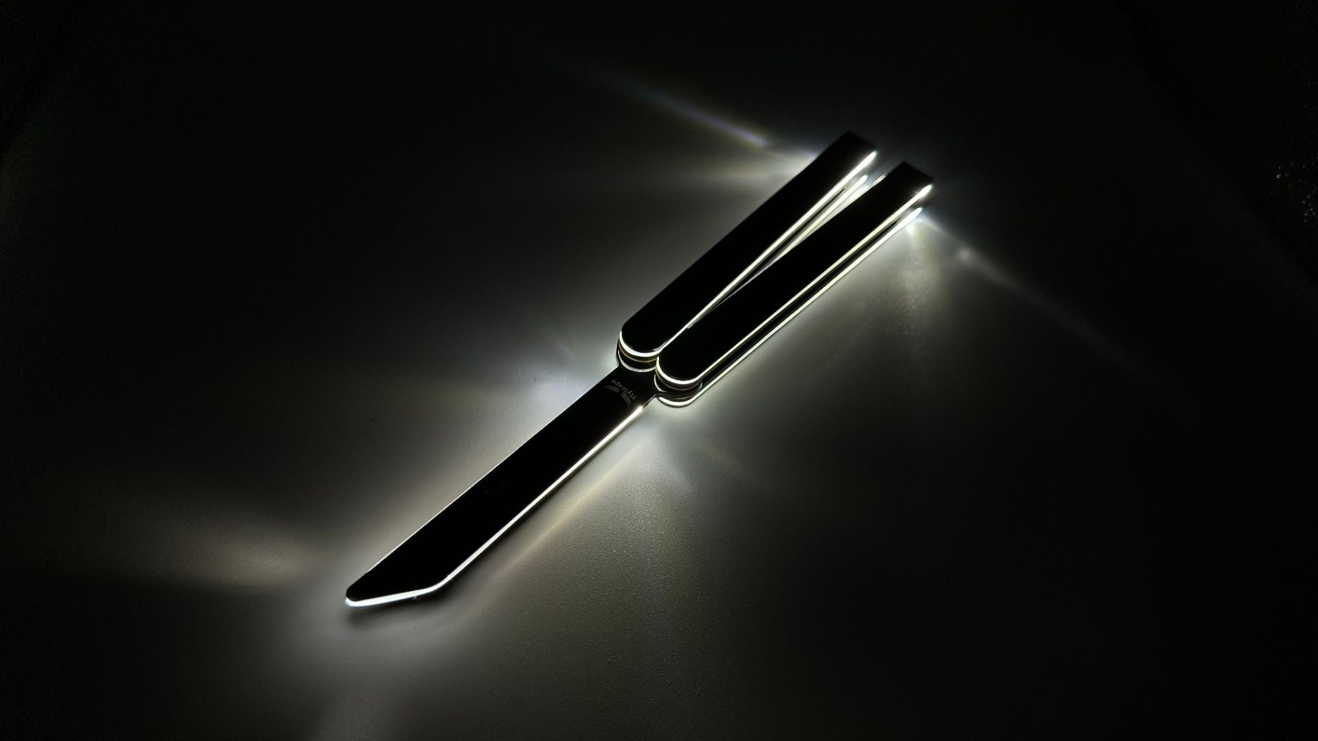 Illuminated balisong trainer with glowing edges, displayed open on a dark surface, casting bright light reflections around its outline.