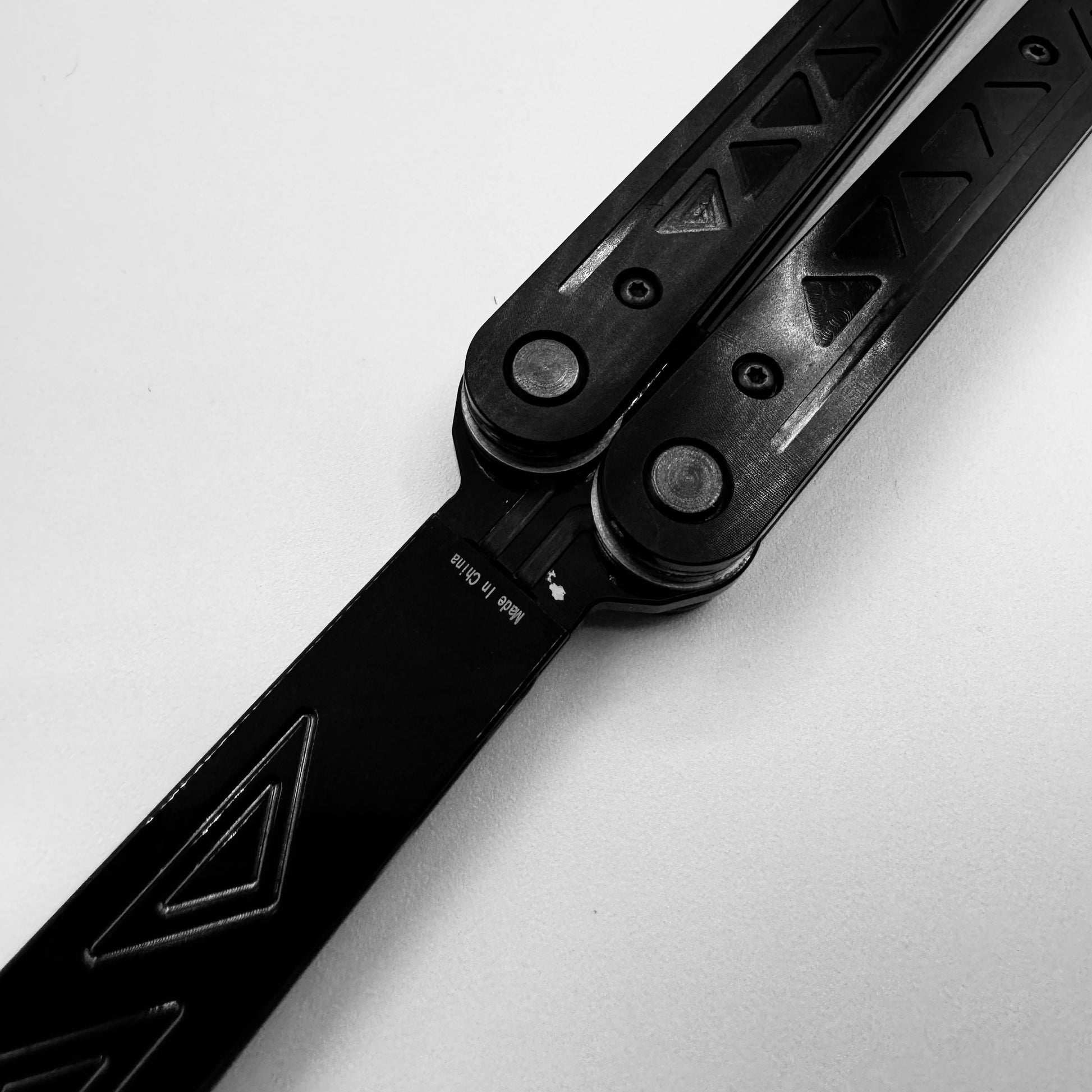 A black and white close-up of a butterfly knife, also known as a balisong or butterfly trainer, with engraved patterns on the blade and textured handles. The knife, also referred to as a Sabre, is shown in an open position against a light background, highlighting its intricate design details and sleek craftsmanship. The lighting emphasizes the contrast between the knife’s dark components and the smooth surface it’s placed on.