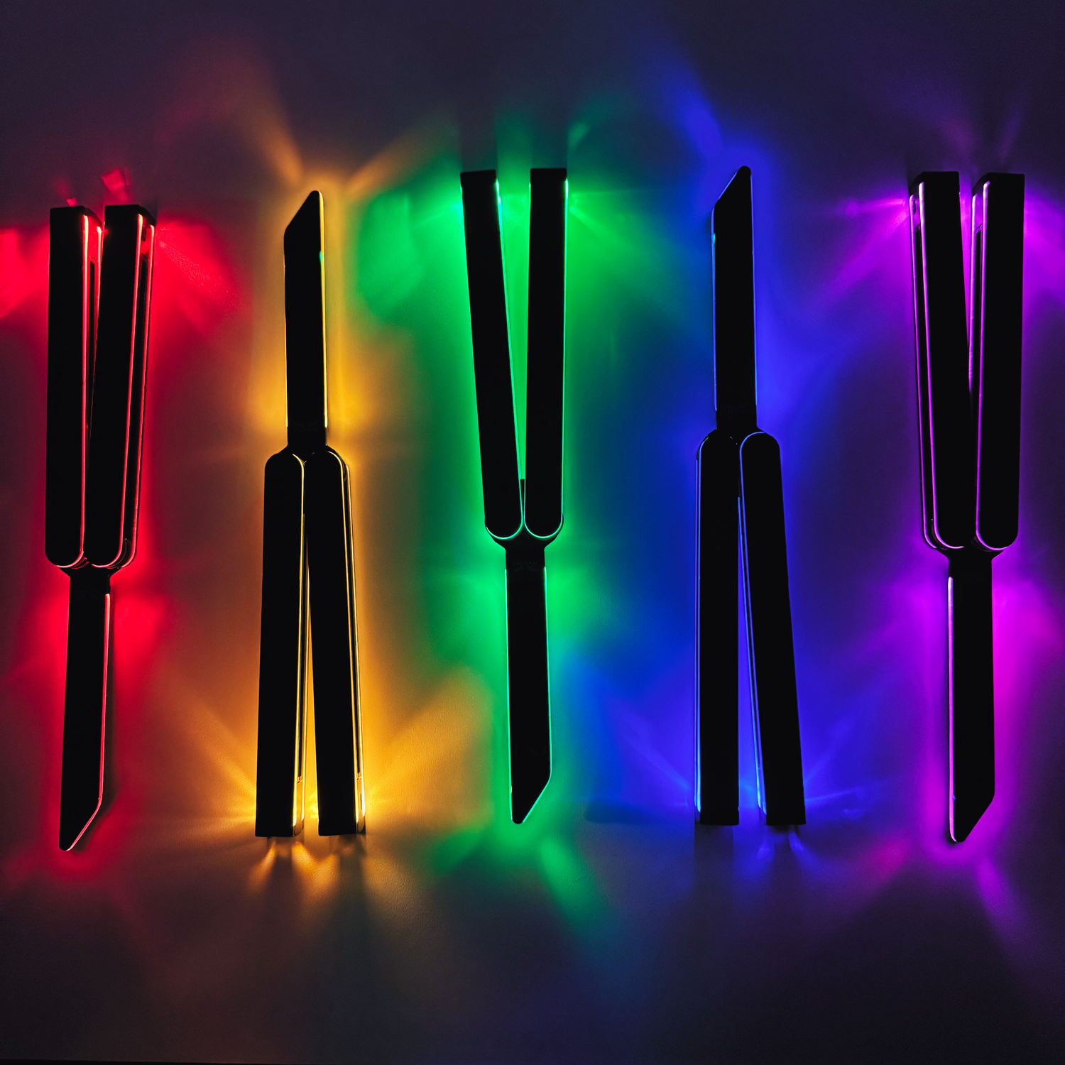Five EOS light-up balisong trainers laid out side by side, each with a colored glow behind it: red, amber/yellow, green, blue, and purple.