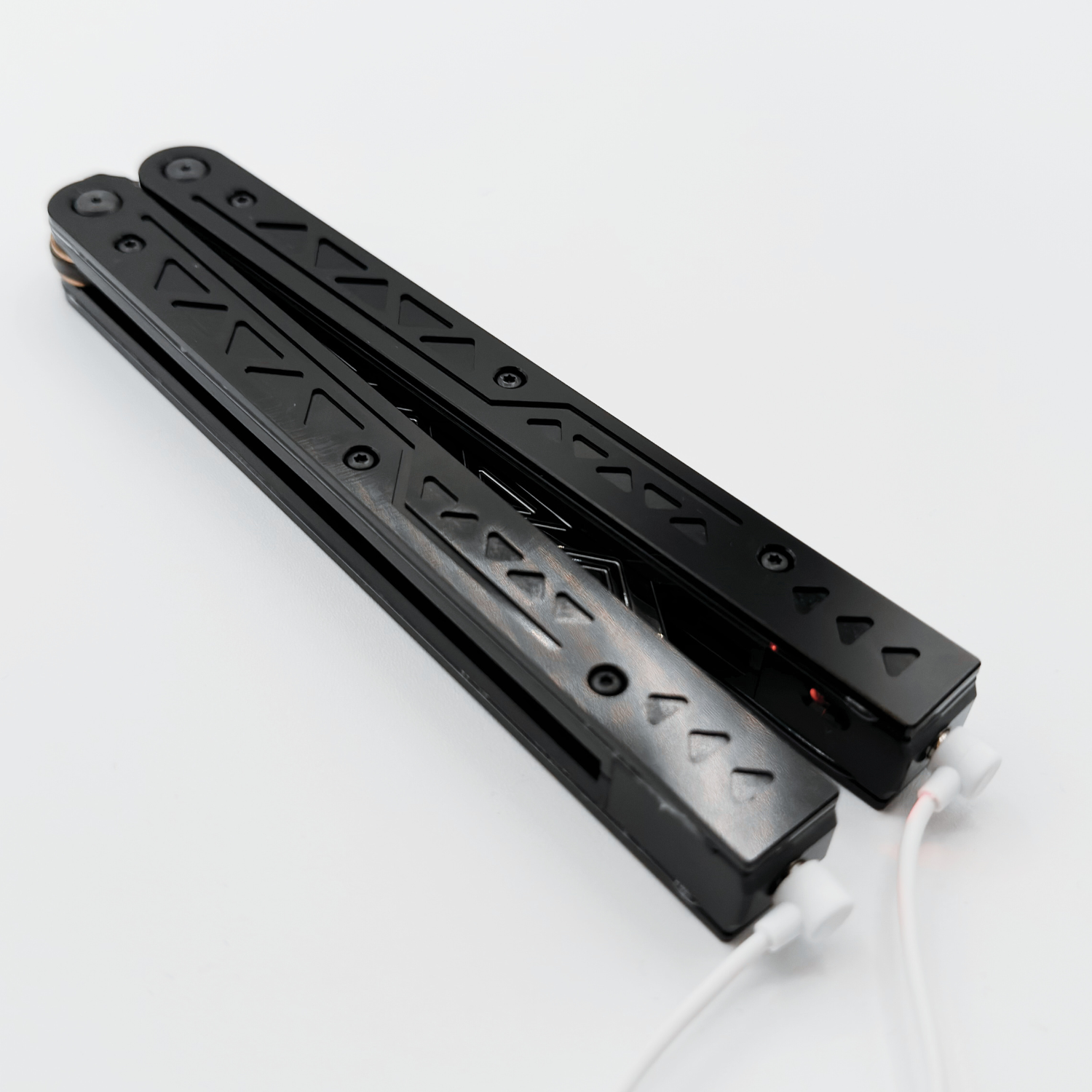 Black balisong trainer with intricate cutout designs on the handles, resting closed on a white background with charging cables attached.