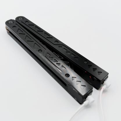 Black balisong trainer with intricate cutout designs on the handles, resting closed on a white background with charging cables attached.