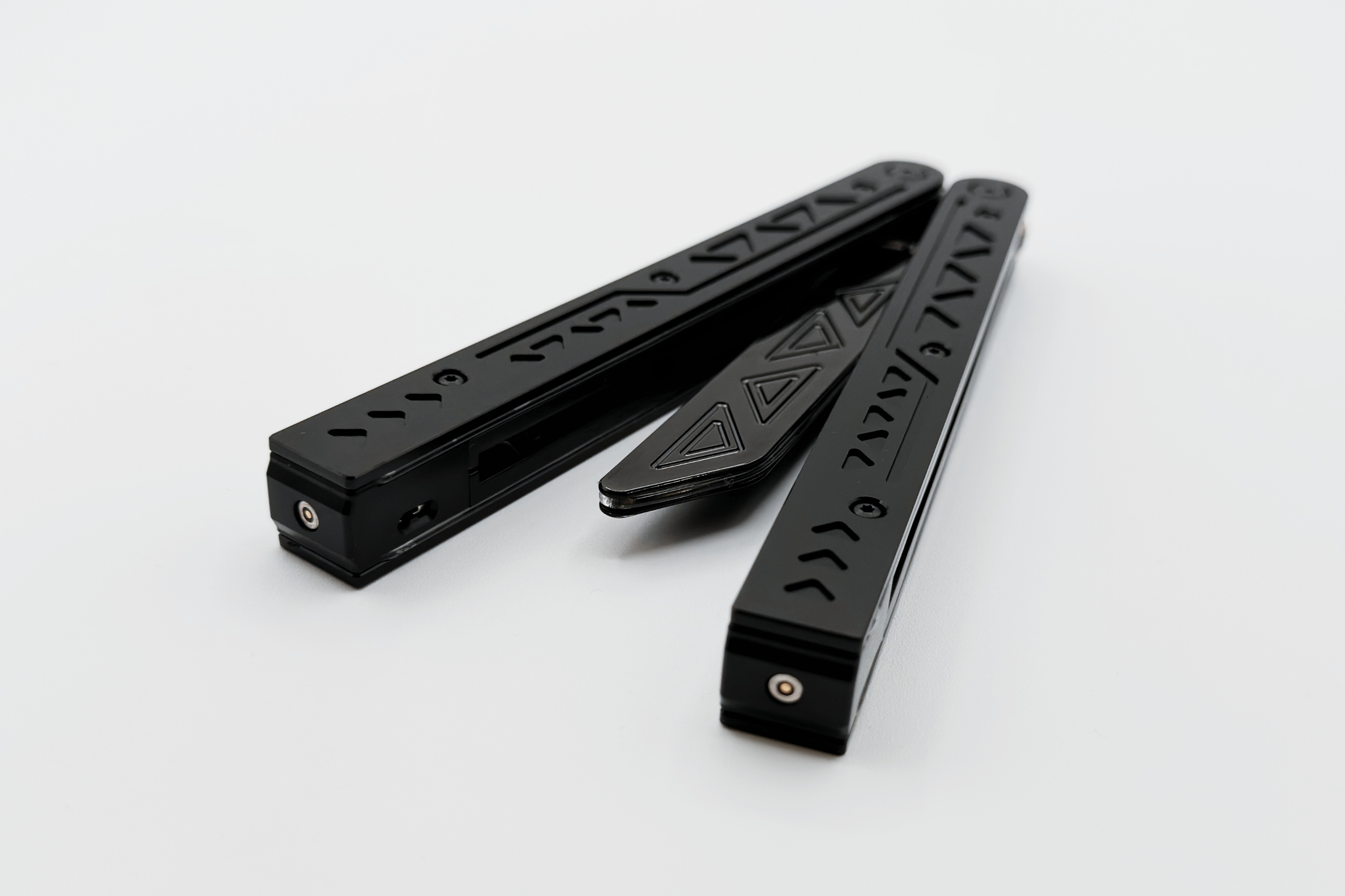 Partially open black EOS balisong trainer with engraved triangular and geometric patterns on the handles, displayed on a white background, highlighting its sleek design and anodized finish.
